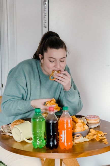 Hungry female in casual clothes eating tasty chips while sitting at wooden table with assorted fast food and bottles of soda
